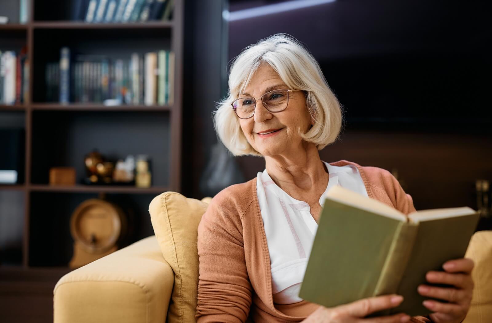 An older adult woman sitting on a couch smiling and holding a book.