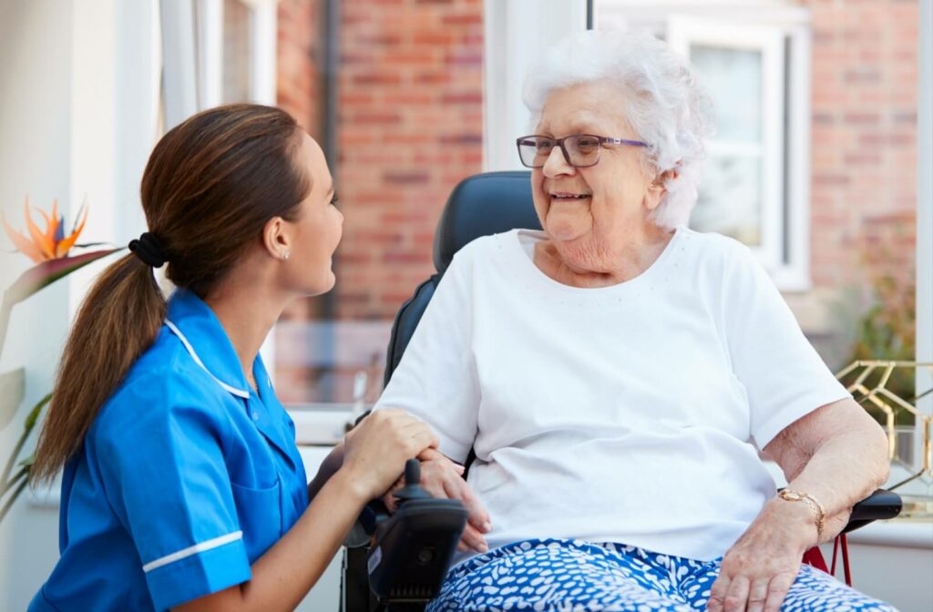 An older adult woman in a senior living facility sitting on a chair smiling and having a conversation with a nurse.