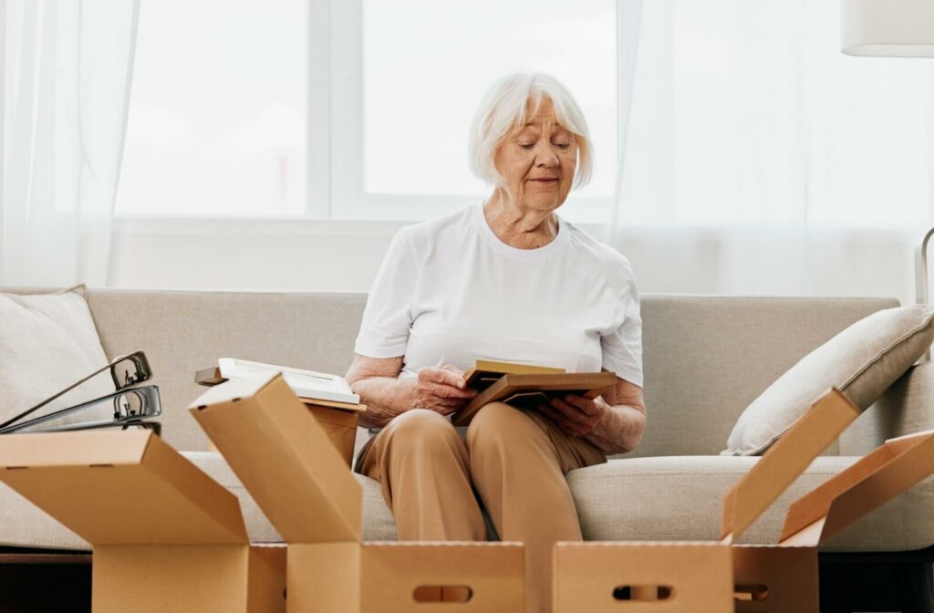 An older woman sitting on a couch holding picture frames and looking at them before packing them into boxes