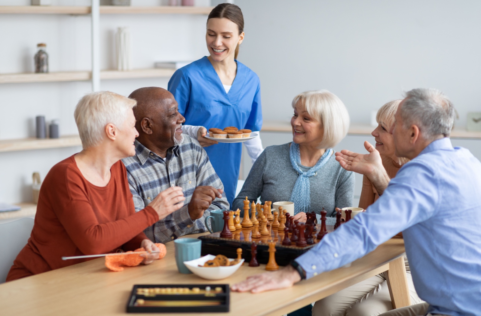 A group of five seniors sitting at a table smiling and playing games while a female nurse stands behind them.