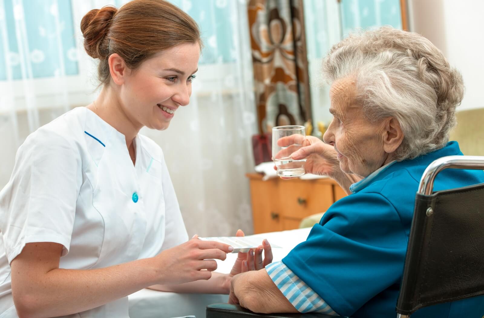 A young female nurse holding a pill case and smiling at an older woman who is holding a glass of water and looking at the nurse.