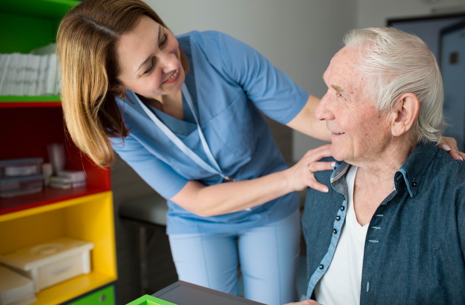 A caretaker touches a senior man on the shoulder, smiling as she helps him in assisted living.