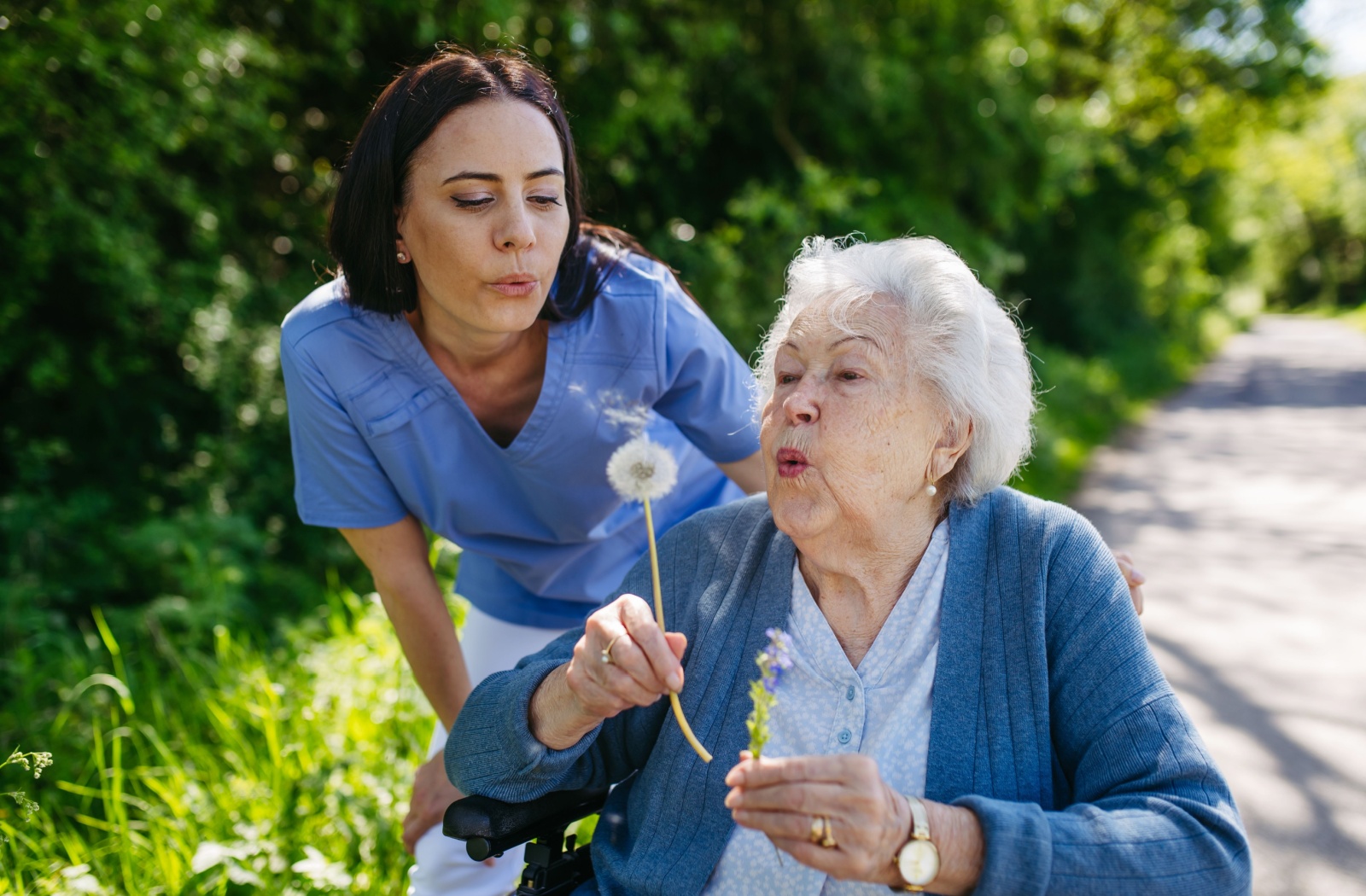 Caregiver enjoying an outdoor moment with an elderly woman in assisted living, both smiling and blowing on dandelions.