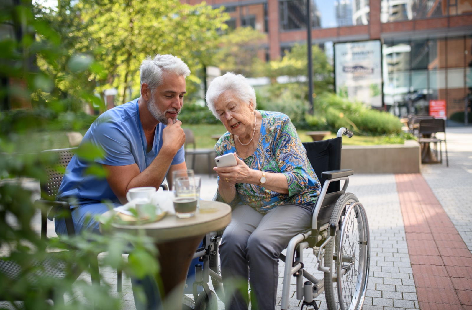 Older adult in a wheelchair showing a phone to a caregiver while sitting at an outdoor café table with drinks