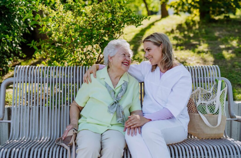 Two people sitting on a bench outside smiling at each other one has an arm around the other in a supportive outdoor setting