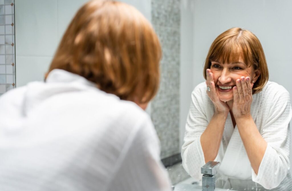 An older adult looks in the bathroom mirror and smiles as they wash their face.