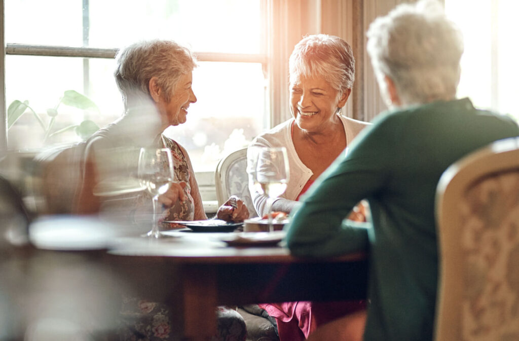 Three older friends sitting around a kitchen table in a sunlit common dining area and smiling.