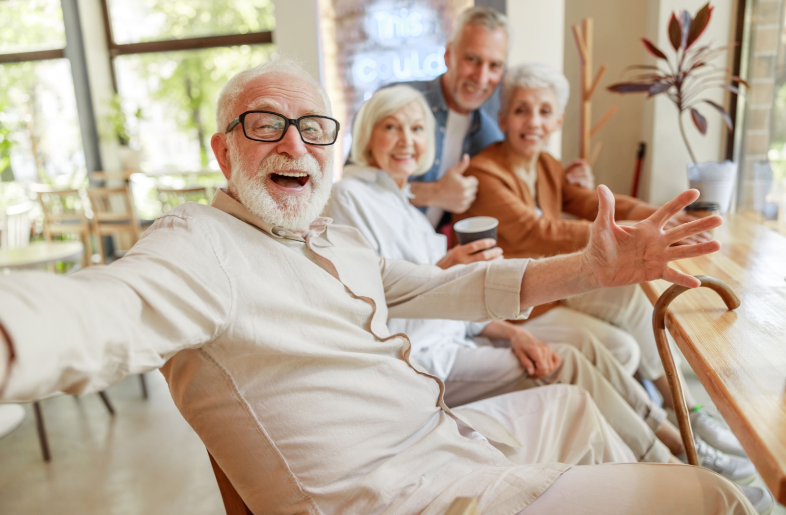 A group of older adults smiling and waving at the camera while sitting in a row of chairs in the lounge of assisted living