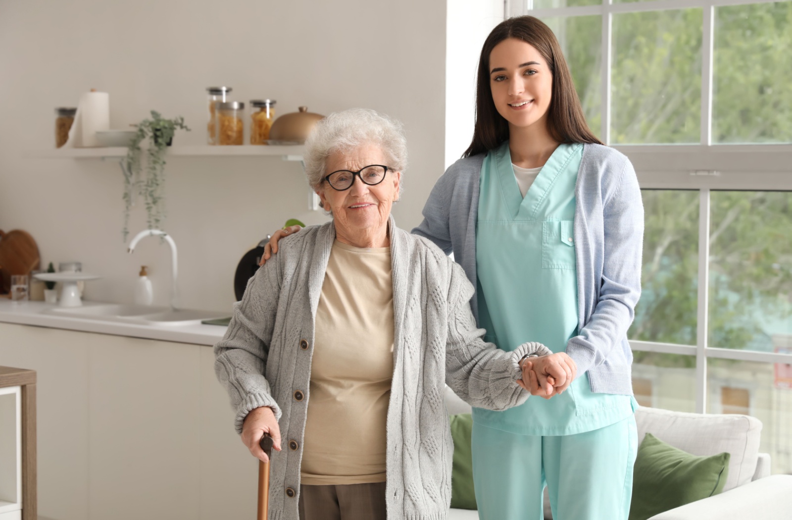 An older adult and a caregiver stand side-by-side in a kitchen in assisted living, holding hands and smiling at the camera