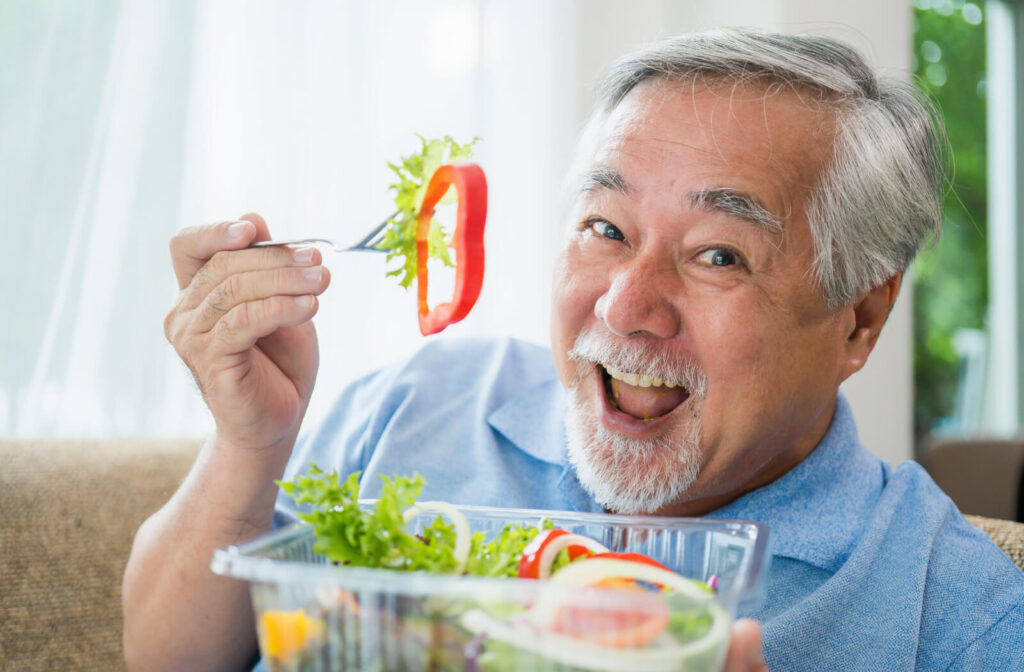 An older adult grins while holding up a sliced red pepper from a salad