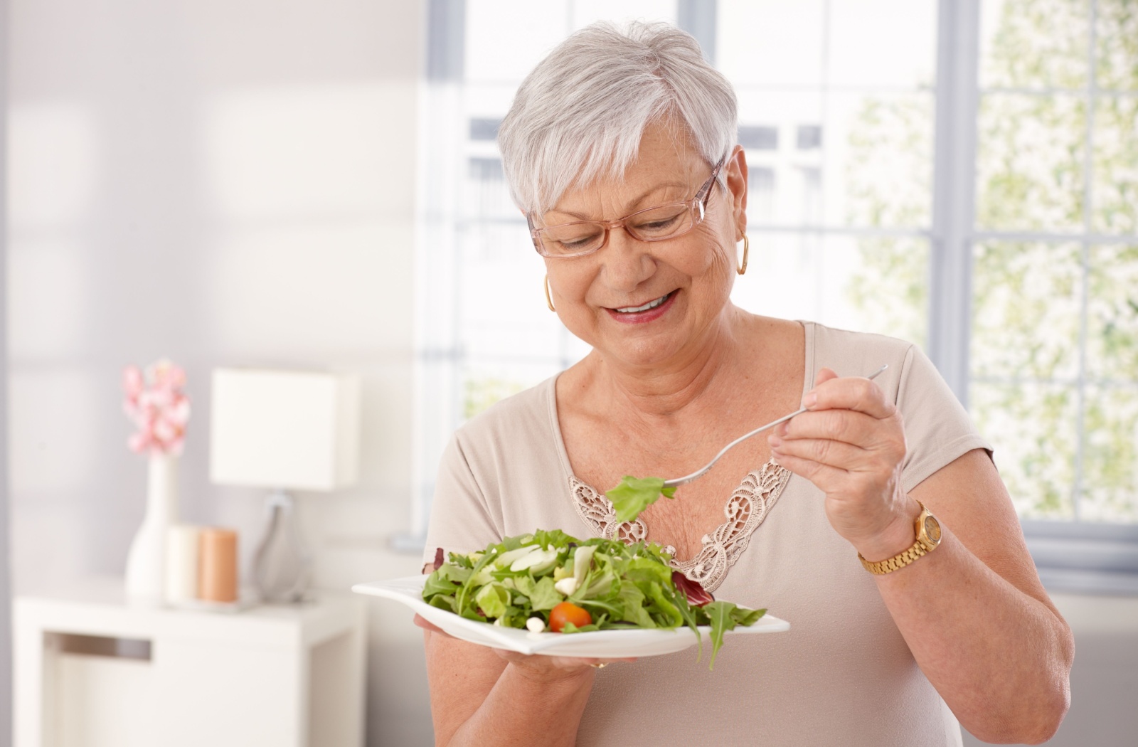 An older adult smiles while holding a bowl of fresh salad in their white kitchen