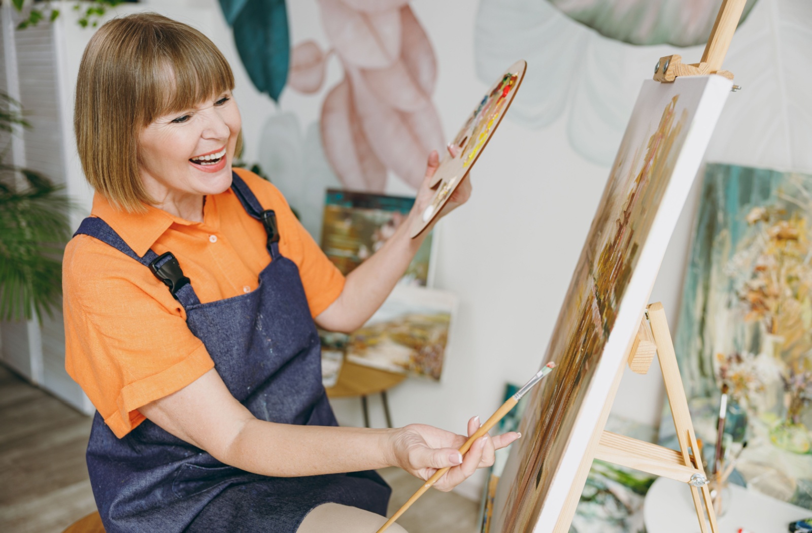 A senior woman painting a canvas with one hand, while holding a color palette in the other.